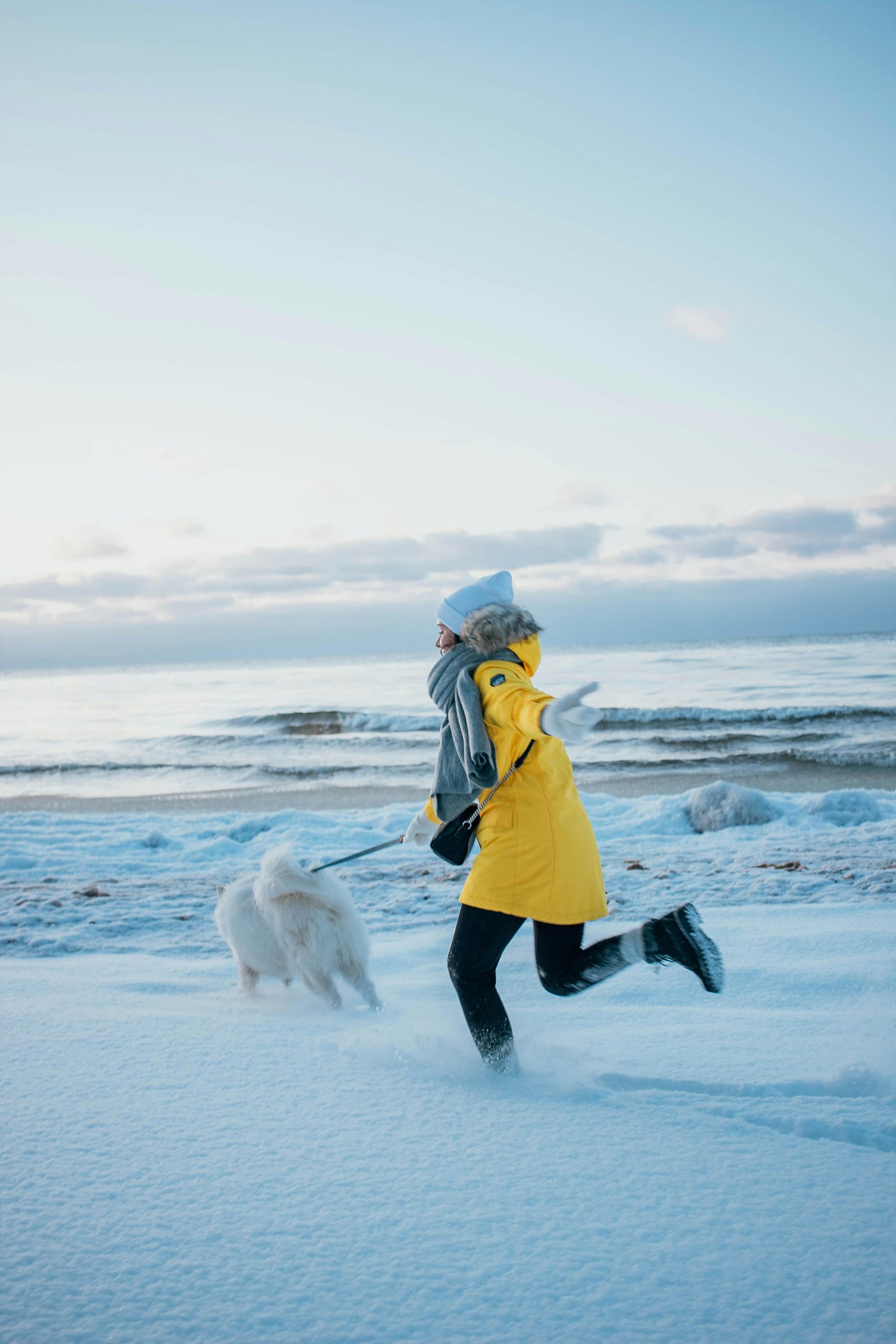 Person in yellow jacket runs with dog by snowy beach during winter in Latvia.